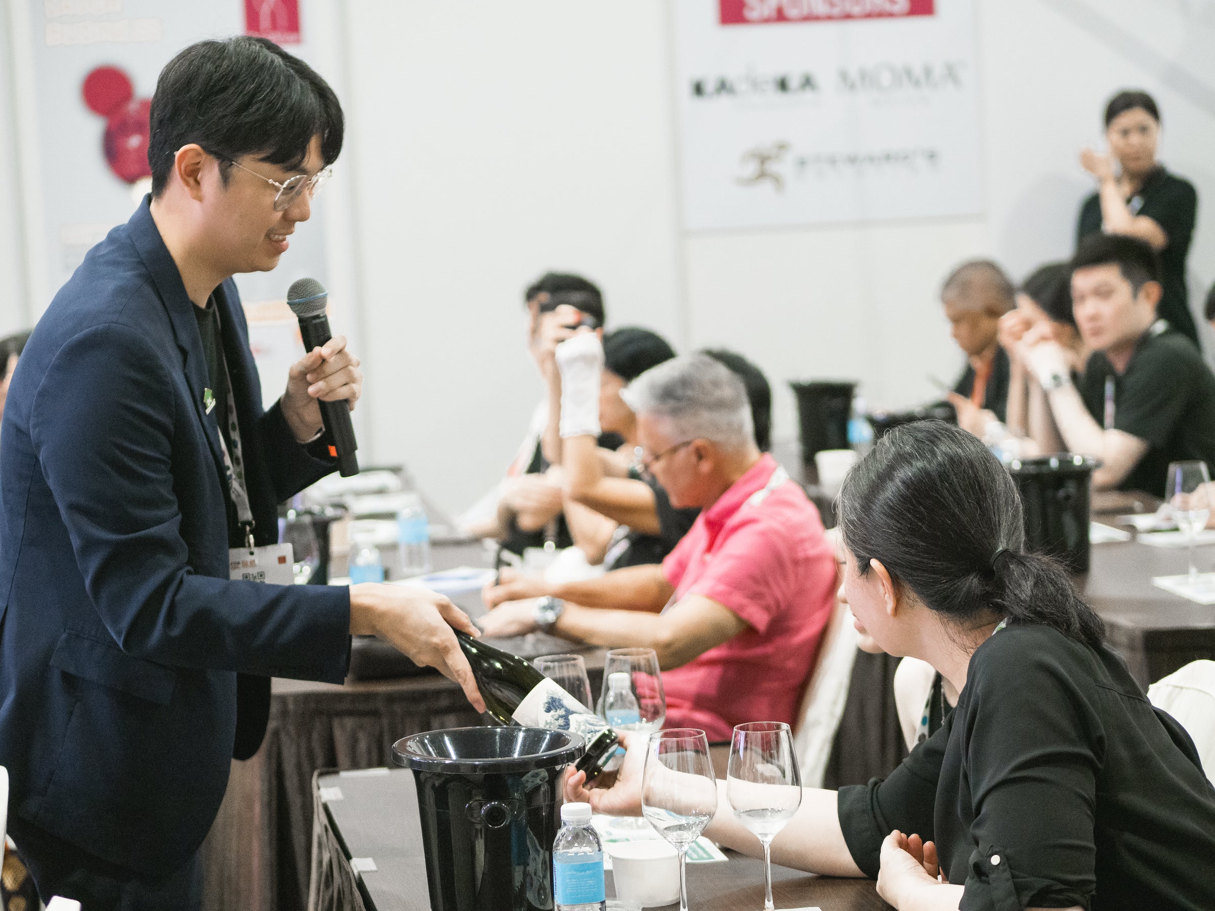 Sake Inn Master Sommelier speaking into a microphone at a conference with attendees listening.