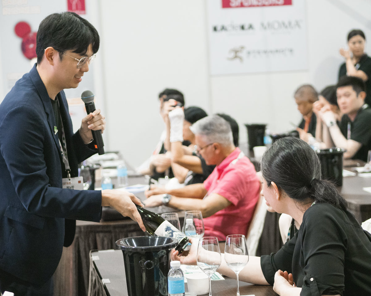 Sake Inn Master Sommelier speaking into a microphone at a conference with attendees listening.
